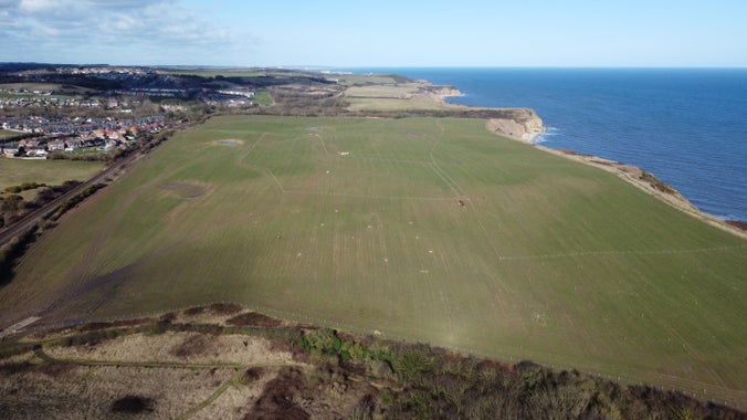 Aerial view of a huge green field on a cliff top on the Durham Coast. The sea to the right is choppy and deep blue with blue sky above. Fields lead into the distance at the top of the image and the houses of Horden and Easington Colliery can be seen on the right.